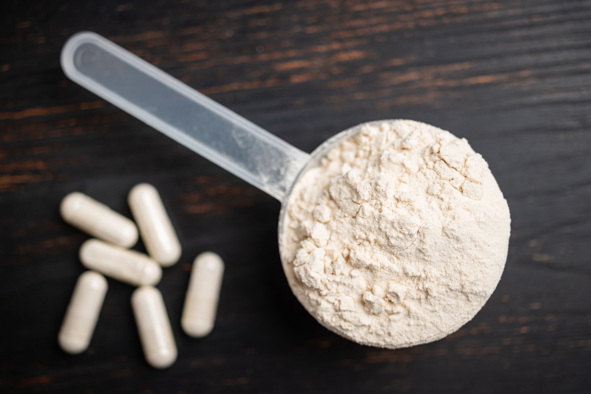 Cucharada de suero de leche o proteína de soja en polvo, cápsulas blancas de aminoácidos, vitaminas y creatina, suplementos alimenticios de culturismo, nutrición deportiva en una tabla de madera oscura, vista superior